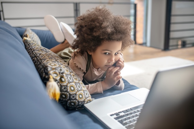 Interessante Information. Interessiertes kleines afroamerikanisches Mädchen mit dunklen lockigen Haaren, das auf den Laptop schaut, der zu Hause auf dem Sofa liegt