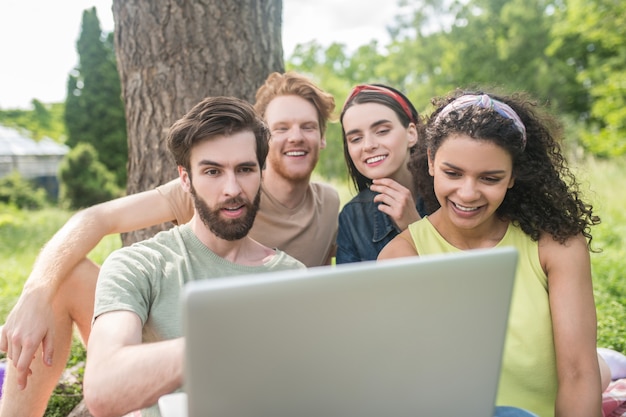 Interessante Information. Vier junge glückliche Freunde, die emotional in den Laptop schauen, verbringen die Sommerfreizeit draußen im Park an einem warmen Tag