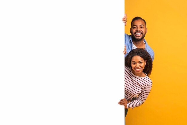 Interessantes Angebot ein glückliches junges schwarzes Paar, das hinter einer weißen Anzeigetafel späht