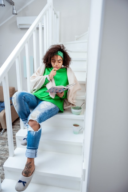 Interessantes Buch lesen. Ungewöhnliche angenehme Frau, die auf Treppen in ihrer Maisonette-Wohnung liegt und Geist mit Buchhandlung beschäftigt