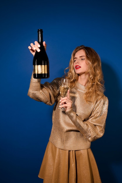 Interesse. Blondes hübsches Mädchen in goldenem Pullover mit Glas, das auf eine Flasche Champagner schaut, die auf Gesichtshöhe auf blauem Hintergrund hält