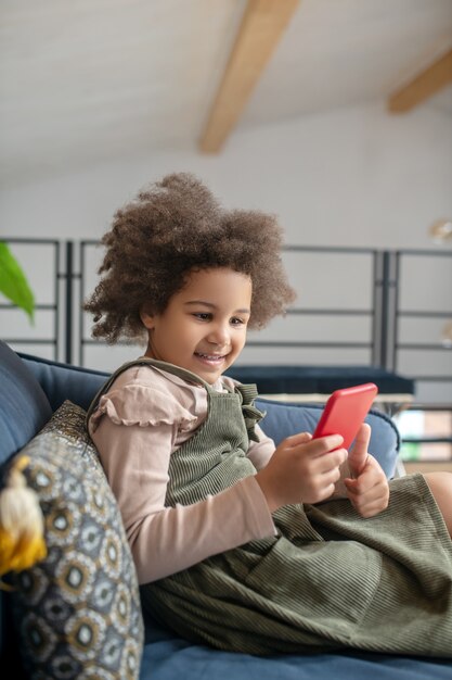 Interesse, Gerät. Süßes kleines dunkelhäutiges Mädchen mit lockigem Haar mit Smartphone, das zu Hause auf einem gemütlichen Sofa sitzt und gut gelaunt ist