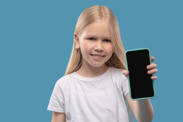 Interesse. Kleines umgängliches lächelndes langhaariges Mädchen, das Smartphone-Bildschirm in der Hand zeigt, der auf hellblauem Hintergrund steht