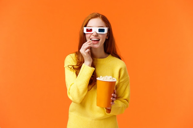 Interessierte und amüsierte, lächelnde geeky rothaarige Frau im gelben Pullover, Film im Kino mit 3D-Brille schauend, Popcorn essen