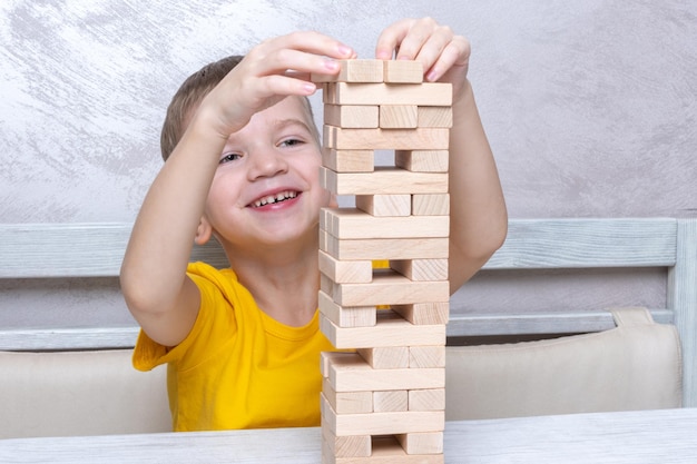 Interessierter glücklicher kleiner blonder Junge, der Brettspiel spielt und Steine vom Holzturm nimmt, um das Gleichgewicht zu halten und Spaß zu haben