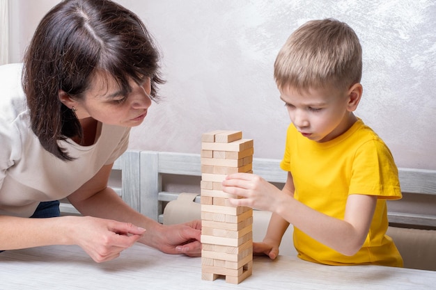 Interessierter glücklicher kleiner blonder Junge spielt Brettspiel mit reifer Mutter mittleren Alters, die Ziegel vom Holzturm nimmt