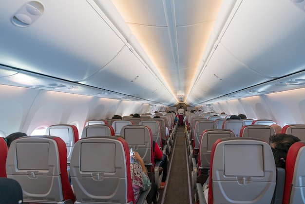 Interior del avión con pasajeros en asientos y azafata en uniforme caminando por el pasillo.