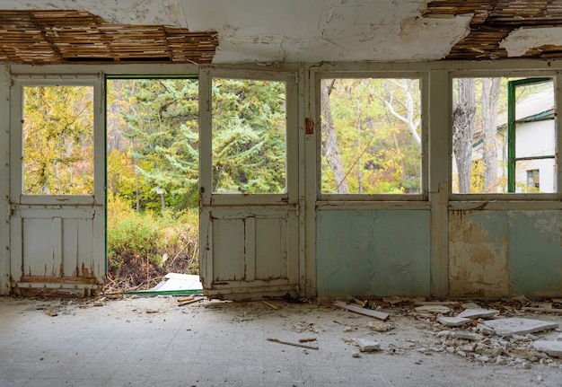 Interior de una casa abandonada. Galería con marcos de puertas y ventanas, vidrios rotos y yeso caído, jardín de otoño cubierto de vegetación que se ve a través de las ventanas