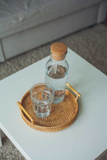 Interior de la casa. Una botella de vidrio con agua, un vaso, una bandeja de mimbre en una mesa de café. Acogedor.