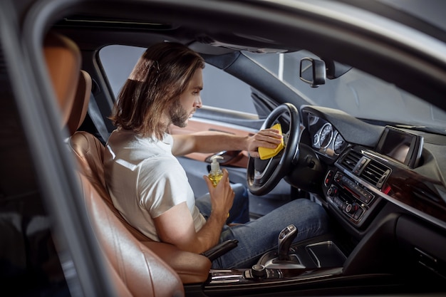 Interior del coche, limpieza. Hombre de pelo largo joven serio concentrado en camiseta y jeans limpiando el volante en el coche con una servilleta