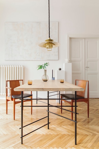 Interior de comedor elegante y moderno con mesa de diseño compartido, sillas, lámpara colgante de oro, cuadros abstractos y accesorios elegantes. Hojas tropicales en florero. Decoración del hogar ecléctica.
