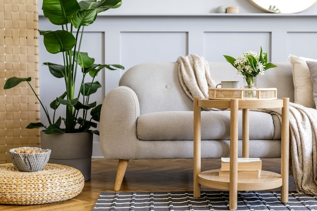 Interior da sala de estar escandinava com sofá cinza de design, mesa de centro de madeira, plantas tropicais, prateleira, espelho, móveis, travesseiro xadrez, bule, livro e elegantes acessórios pessoais na decoração da casa.