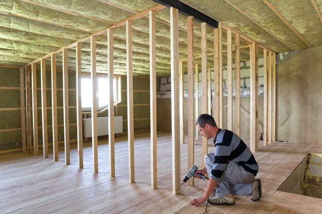 Interior da sala isolada no sótão com piso de carvalho em reconstrução. Jovem trabalhador profissional usa nível e chave de fenda para instalar a moldura de madeira para futuras paredes. Conceito de renovação e melhoria.