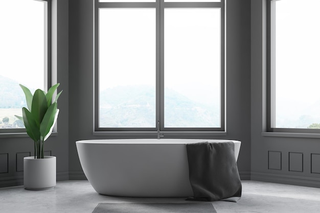 Interior de banheiro minimalista com paredes cinzentas, chão de concreto, grandes janelas e banheira branca com toalha cinzenta.