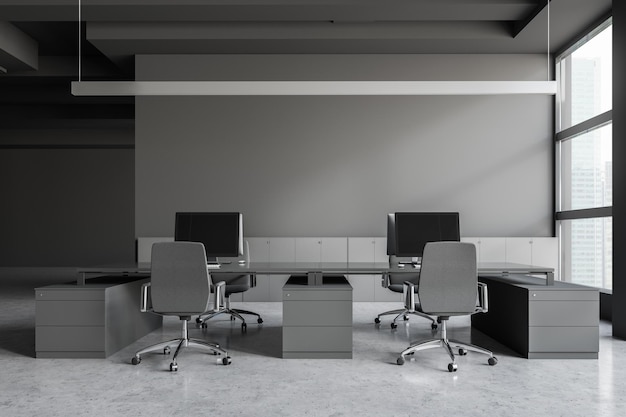Interior de escritório cinza com janelas panorâmicas, paredes cinzentas, chão de concreto, mesas de computador cinzentas maciças com cadeiras e armários perto da parede.