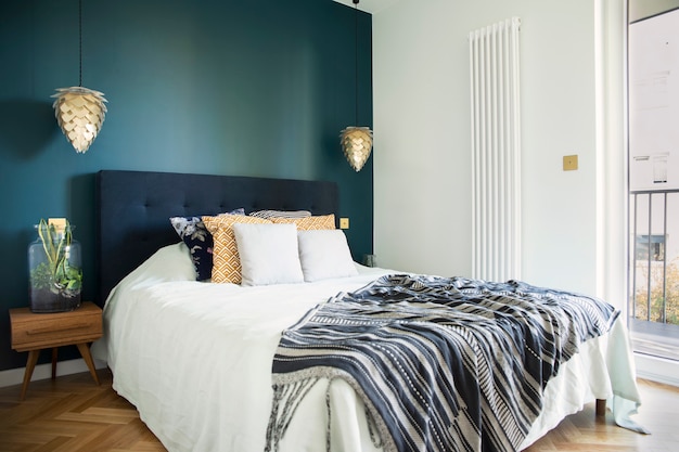 Interior de dormitorio soleado elegante y moderno con mesita de noche de madera, jardín en un frasco, ropa de cama blanca, almohadas de colores y manta. Espacio con paredes azules y parquet de madera marrón.