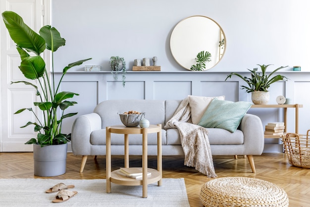 Interior de la elegante sala de estar con sofá gris de diseño, mesa de café, puf, canasta, estante, espejo, plantas tropicales, decoración, alfombra, almohadas y elegantes accesorios personales en la decoración del hogar moderno.