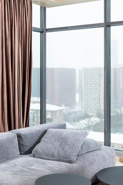 El interior de una elegante sala de estar con ventanas panorámicas en un edificio de gran altura de la ciudad. Acogedor sofá gris y cortinas marrones. Vertical.