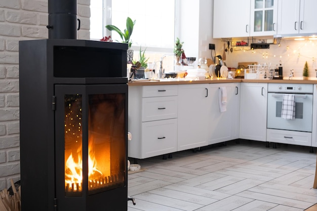 El interior festivo de la casa está decorado para Navidad y Año Nuevo en estilo loft con chimenea de estufa negra Árbol de Navidad Cálida sala de estudio con cocina blanca que quema madera acogedora y calefacción de hogar