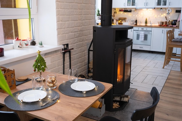 El interior festivo de la casa está decorado para Navidad y Año Nuevo en estilo loft con chimenea de estufa negra Árbol de Navidad Cálida sala de estudio con cocina blanca que quema madera acogedora y calefacción de hogar