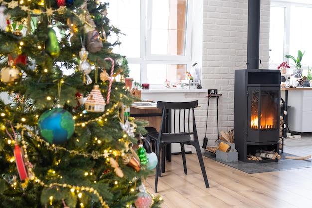 El interior festivo de la casa está decorado para Navidad y Año Nuevo en estilo loft con chimenea de estufa negra Árbol de Navidad Cálida sala de estudio con cocina blanca que quema madera acogedora y calefacción de hogar