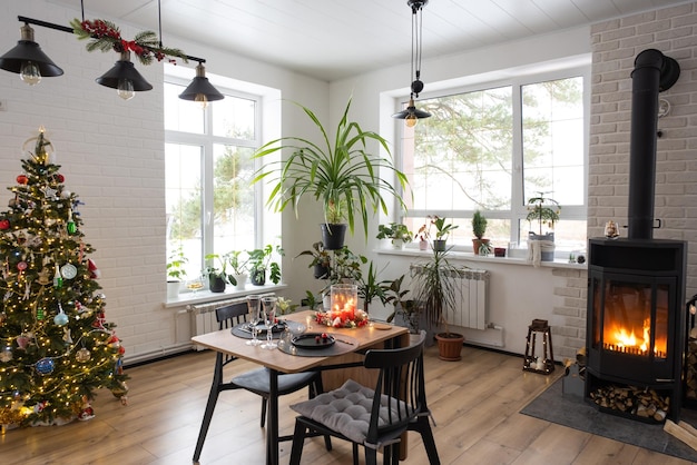 El interior festivo de la casa está decorado para Navidad y Año Nuevo en estilo loft con chimenea de estufa negra Árbol de Navidad Cálida sala de estudio con mesa puesta quemando madera acogedora y calefacción de hogar