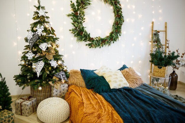 Interior festivo de Natal nas cores cinza e branco. Uma coroa de flores verde redonda está pendurada na parede. Uma cama coberta com cobertores aconchegantes fica em uma casa decorada com uma guirlanda.