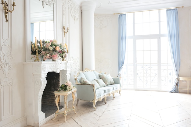 Interior luminoso de lujo en estilo barroco. Una habitación espaciosa con un hermoso mobiliario de carretera elegante, una chimenea y flores. Planta de estuco en las paredes y parquet de madera clara.