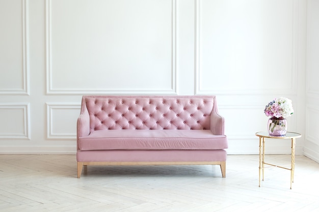 Interior minimalista moderna sala de estar com um sofá rosa e uma mesa com um vaso de buquê de flores contra a parede branca. a espaçosa sala de estar de estilo clássico possui um sofá de veludo. conceito de conforto