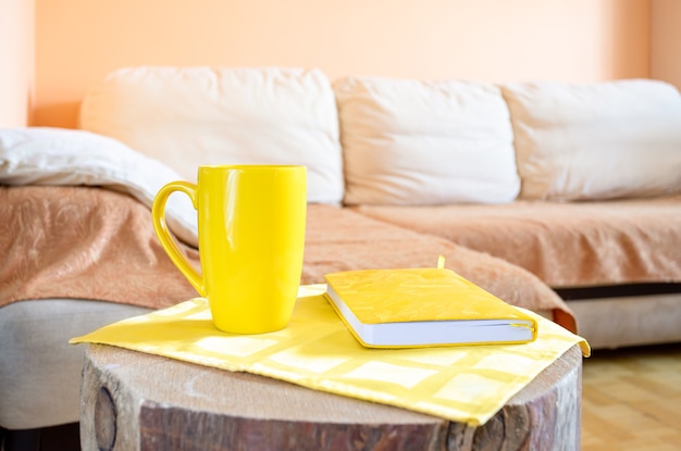 Interior moderno e aconchegante de casa minimalista para relaxar: confortável sofá de canto grande leve, coto de madeira criativo como mesa com copo amarelo e bloco de notas close-up.