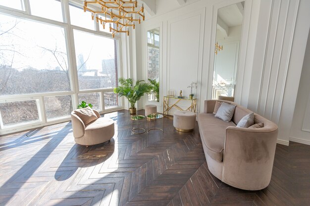 Interior muito luminoso e luminoso de uma sala de estar acolhedora e luxuosa com elegantes móveis bege suave com elementos metálicos dourados, janela enorme para o chão e parquet de madeira