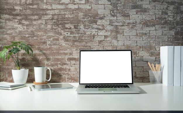 Interior de oficina en casa con computadora portátil, libros, taza de café y planta de interior en mesa blanca