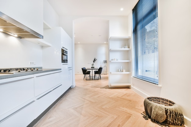 Interior de la sala de la cocina de la casa con gabinetes minimalistas blancos y electrodomésticos empotrados y paso abierto al comedor con mesa