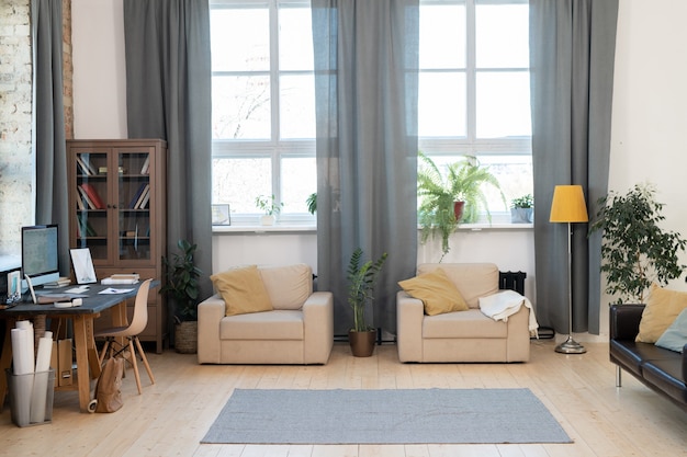 Interior de sala de estar en casa o apartamento moderno con dos sillones, alfombra en el piso, sofá, mesa con computadora y gabinete con libros