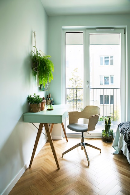 Interior scanidinavian moderno de escritório em casa com mesa de design, poltrona, acessórios de escritório, janela grande e plantas. Espaço elegante e ensolarado com parquet marrom de madeira