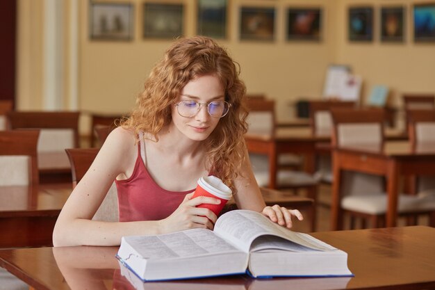 El interior de la tienda de la mujer estudiante que está en la biblioteca, leyendo un libro y tomando café, pasando tiempo en la biblioteca, preparándose para las clases, una mujer vestida con una camiseta marrón, parece concentrada. Concepto de educación