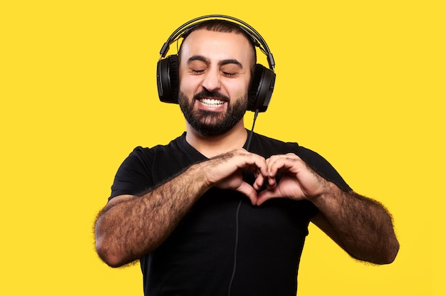 Internationaler DJ Day. Junger Mann mit Bart und Kopfhörern, die Herz mit Händen zeigen und gelb lächeln