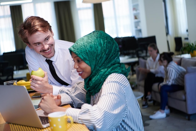 internationales multikulturelles geschäftsteam. mann, der apfel isst, afrikanische muslimische frau, die grünen hijab trägt und tee trinkt, während sie gemeinsam am laptop im entspannungsbereich bei modernen offenen startu arbeiten