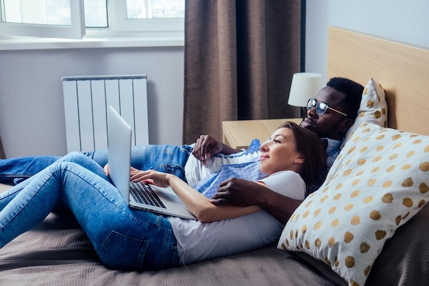 Internationales Paar lächelt und schaut auf den Laptop, während es sich zu Hause auf dem Bett im Schlafzimmer ausruht