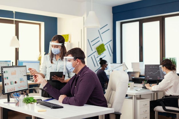 Internationales Team, das Finanzanalysen mit Gesichtsmaske als Sicherheitsvorkehrung während der globalen Pandemie durchführt. Multiethnisches Team in einem neuen normalen Finanzbüro in einem Unternehmensunternehmen, das auf dem Computer tippt, taki