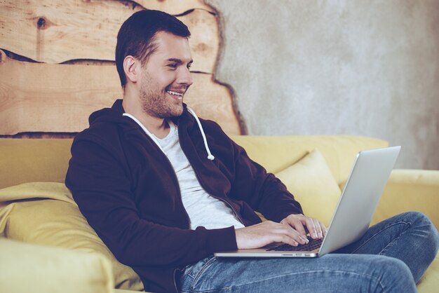 Internet de navegação em casa. Jovem alegre usando seu laptop com um sorriso enquanto está sentado no sofá em casa