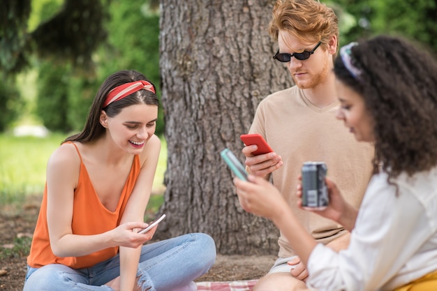 Internet. Jovem interessado em óculos de sol e garotas sorridentes, sentado olhando para smartphones em um piquenique no ar quente