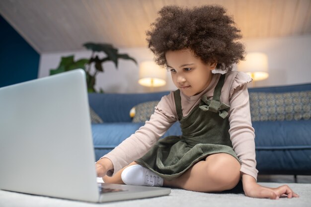 En Internet. Niña afroamericana de pelo rizado sentada cerca de la computadora portátil en el piso en casa, interesada enfocada