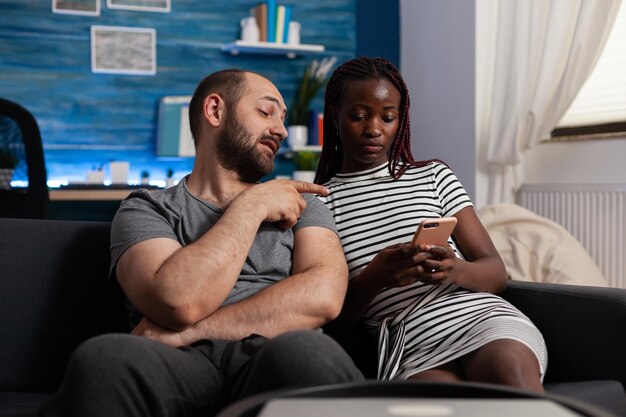 Interracial Menschen, die Technologie für die Unterhaltung zu Hause verwenden. Afroamerikanerfrau, die modernes Smartphone hält, während kaukasischer Mann digitales Gerät betrachtet. Multiethnisches Paar