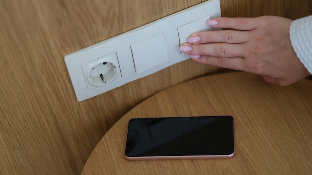 Interruptor de luz de mano en la pared de la mujer y primer plano del teléfono inteligente