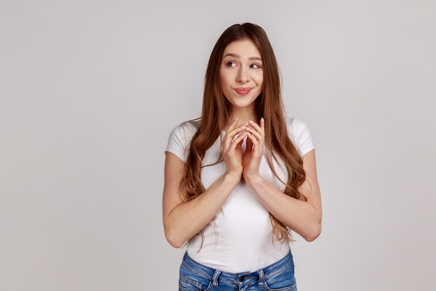 Intrigando a una mujer atractiva y tortuosa agarrándose de las manos y pensando en un plan complicado que tiene una idea astuta y astuta para bromear usando una camiseta blanca Foto de estudio en interiores aislada en un fondo gris