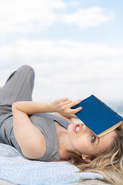 Foto intrigante mujer rubia sosteniendo un libro mientras se encuentra en la playa y cubriéndose la cara