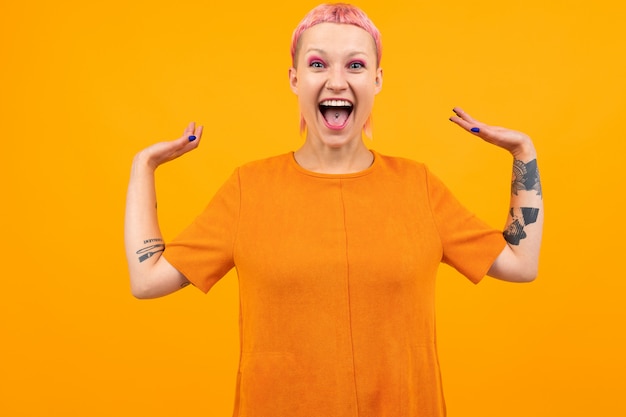 Inusual mujer bonita con cabello rosado corto y tatuaje grita de felicidad aislado en naranja