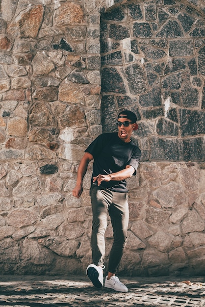 Inventando su propio estilo de baile. Longitud total de apuesto joven africano en ropa casual haciendo una mueca mientras se mueve y gesticula contra la pared de piedra al aire libre