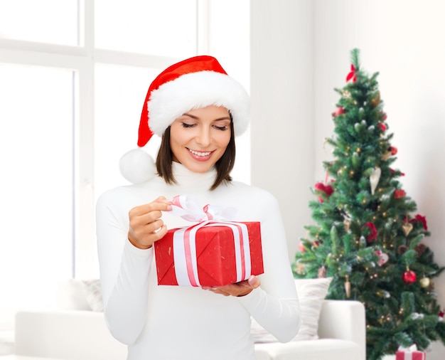 inverno, felicidade, feriados e conceito de pessoas - mulher sorridente com chapéu de ajudante de papai noel com caixa de presente sobre a sala de estar e o fundo da árvore de natal